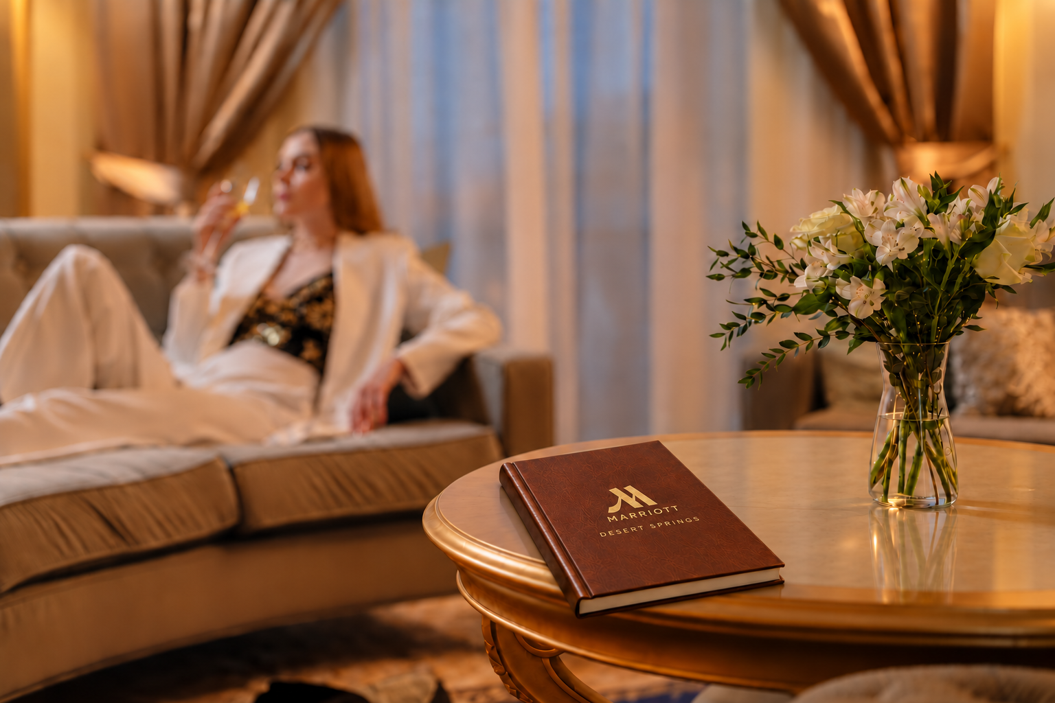A leather-bound hotel book on a gilded table beside white flowers, with a guest relaxing on the sofa behind